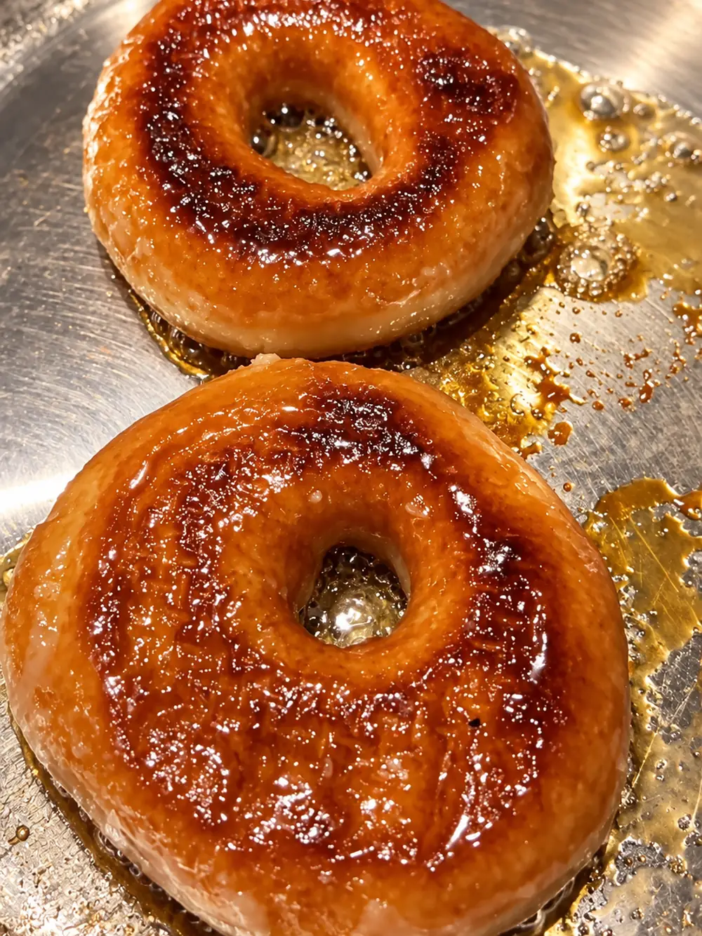 Two glazed donuts caramelising in a stainless steel skillet with melted amber sugar around them