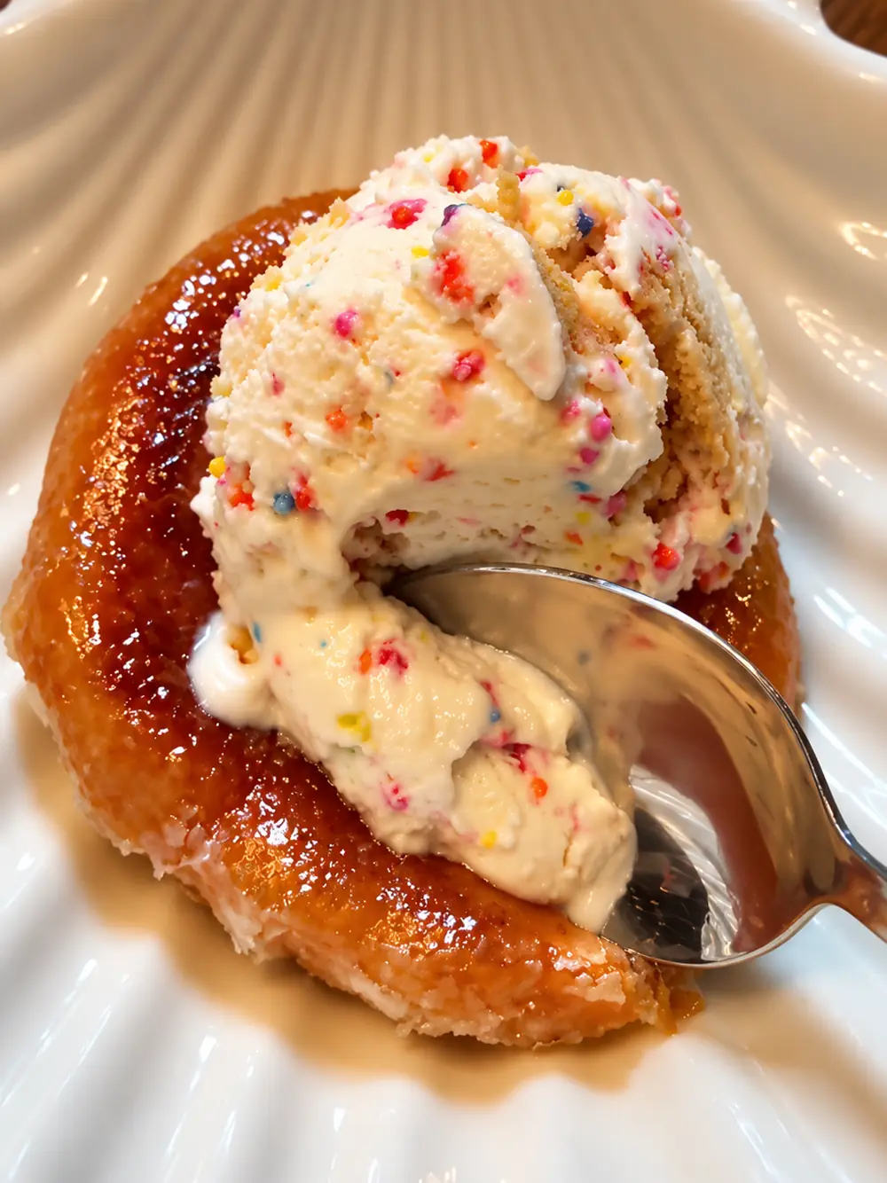 A spoon breaking through funfetti ice cream into the soft interior of a caramelised glazed donut