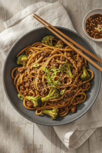 Ground beef and broccoli noodles in a wide matte charcoal ceramic bowl, glossy brown sauce coating thick lo mein noodles